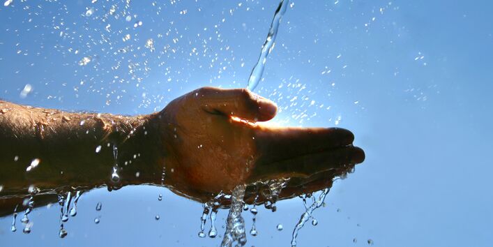 Zwei Hände werden in einen Wasserstrahl gehalten. Im Hintergrund scheint die Sonne