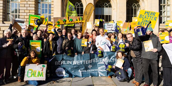 Gruppenfoto einer großen, jubelnden Menschenmenge. Sie halten Schilder, blaue Gießkannen und ein Banner mit der Aufschrift ‚BERLIN WETTERFEST MACHEN‘ und dem Logo ‚BAUM ENTSCHEID BERLIN‘ hoch. Die Stimmung ist feierlich.