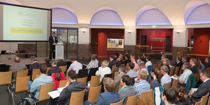 Viele Personen sitzen vor einer Bühne und hören einem Redner zu