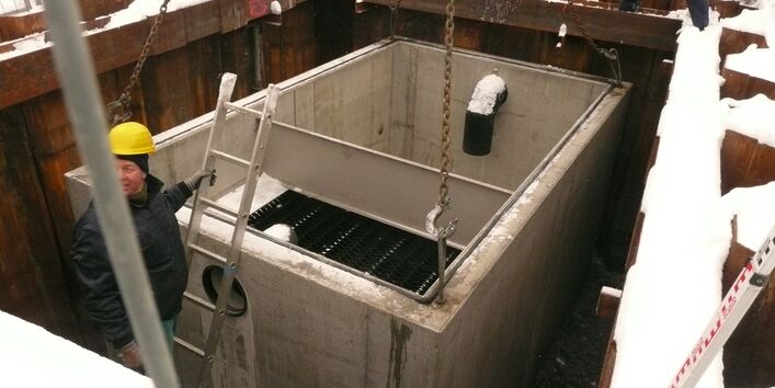 Ein vierreckiges Betongebilde ist der Regenwasserauslass mit Gitter und Rohabfluss hängt an vier Kettenhaken in einer Grube. Ein Mann mit gelben Helm steht dort auf der Leiter. Schnee liegt verteilt überall.