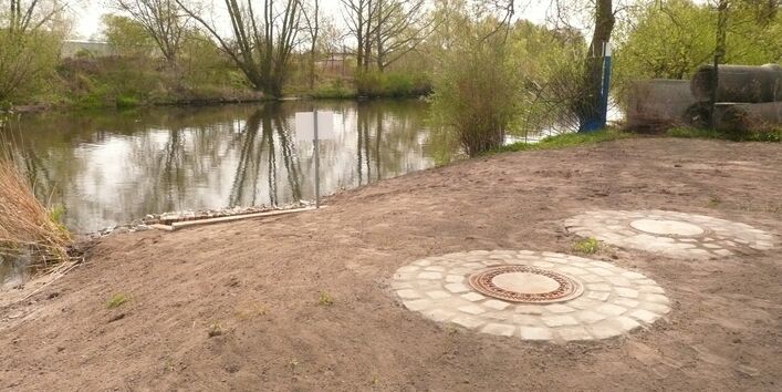 Oberirdisch wird der Regenwasserauslass durch Schachtdeckel abgedeckt. Die Schachtdeckel sind von Pflastersteinen umrahmt. Der Blick geht auf Wasser, die Ränder sind mit Bäumen und Gebüsch bewachsen.