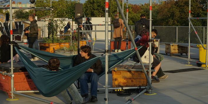 Auf einer Dachterrasse sitzen Menschen auf Metallboxen und in einer Hängematte. Rundherum Pflanzkübel und Gerüstelemente.