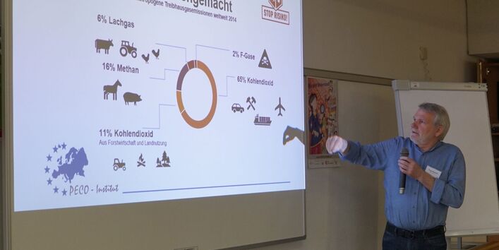 Vor einer Leinwand, auf die eine Präsentationsfolie projiziert wird, steht eine Person mit Mikrofon, die etwas erläutert und auf die Leinwand deutet.