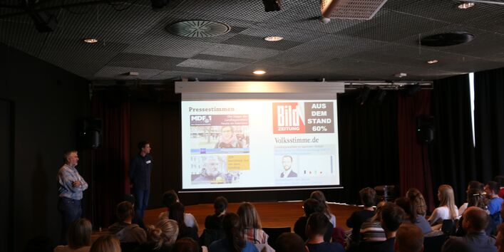 Fotografie einer Präsentation aus der Perspektive eines Zuhörenden. Neben einer Power-Point-Präsentation stehen zwei Vortragende. Im Vordergrund sind mehrere sitzende Personen zu erkennen.