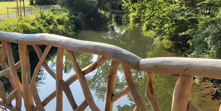 Fotografie von einer Holzbrücke auf einen darunterliegenden Bach neben einem Weg im Grünen.