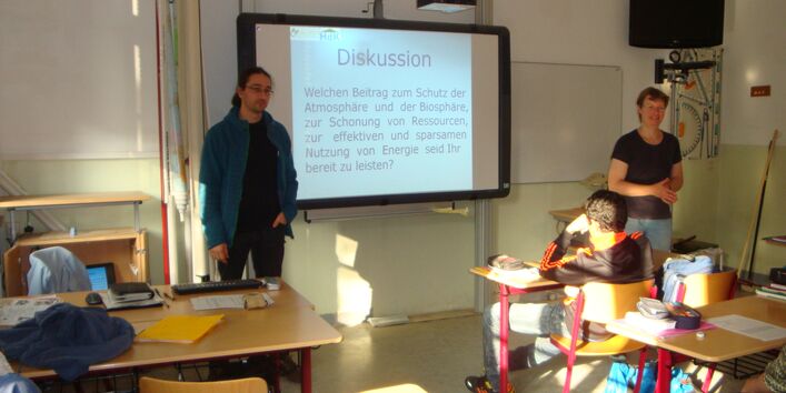 Zwei Seminarleiter stehen vorne im Klassenraum und diskutieren mit der Klasse.