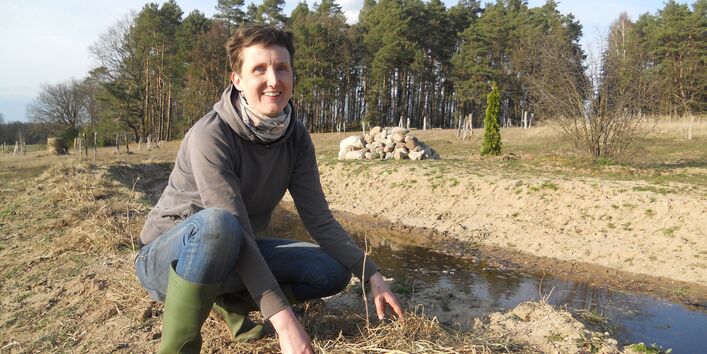 Eine Person in Gummistiefeln hockt neben dem Wassergraben.