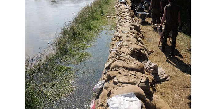 Fotografie eines Sandsackwalls. Links des Walls ist Wasser, aus dem überschwemmte Gräser herausragen, rechts gehen und stehen einige Personen mit Schubkarren auf einem Weg.