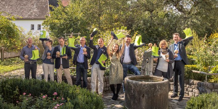 Absolvent/-innen der Weiterbildung stehen locker im Garten hinter einem runden Brunnen und halten jeder ein Paar hellgrüne Gummistiefel und eine hölzerne pokalähnliche Form in den Händen, die sie fröhlich zeigend hoch halten. Im Hintergrund sieht man belaubte Bäume, Sträucher Hausdächer sowie einen Gartenzaun am Rand.
