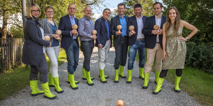 Teilnehmer/-innen der Pilotschulung stehen im Halbkreis auf einem Kiesweg vor blauem Himmel und belaubten Bäumen. Sie halten ein hölzerne pokalähnliche Form in Händen, eine Form steht auf dem Boden vor dem Halbkreis. Die Teilnehmer/-innen tragen hellgründe Gummistiefel.