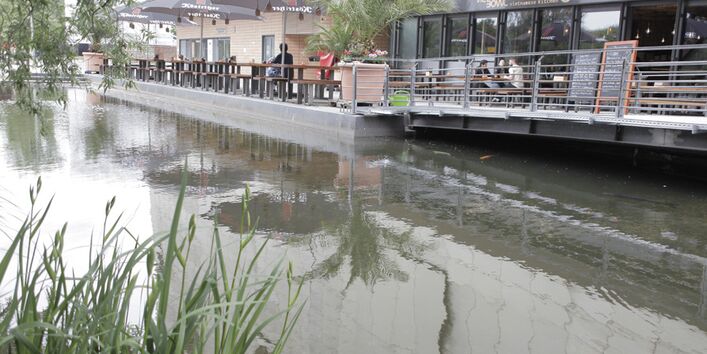 Im Vordergrund fließt ein Fluss. Im Hintergrund sitzen mehrere Menschen an Tischen eines Restaurants, dessen Terrasse über den Fluss ragt.