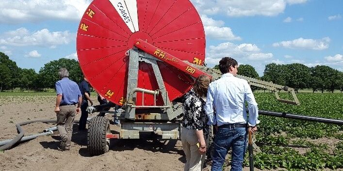 Exkursionsteilnehmende betrachten eine landwirtschaftlich genutzte Beregnungsfläche. Das Beregnungsgerät erscheint als große runde rotes Gebilde im Hintergrund vor wolkigem Himmel.