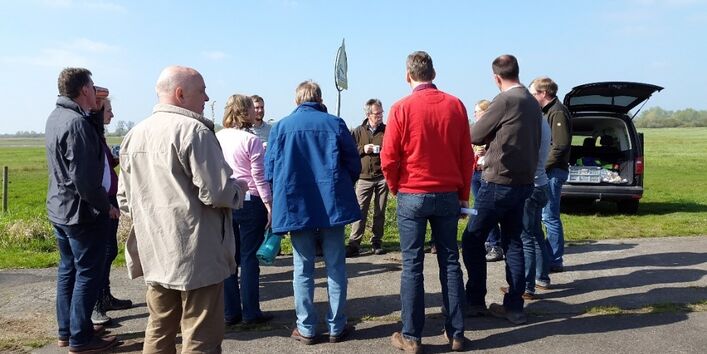 Personen stehen in Gruppe mit den Rücken zum Betrachter auf einem Teerweg. Rechts von der Gruppe steht ein Auto mit geöffneter Kofferraumklappe. Im Hintergrund sind Wiese und blauer Himmel zu sehen.