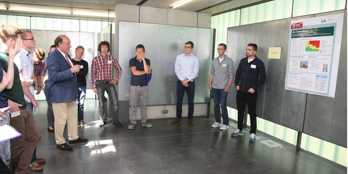 Studierende stehen kreisförmig um einen Dozenten. Im Hintergrund befinden sich silbernfarbene Stellwände.