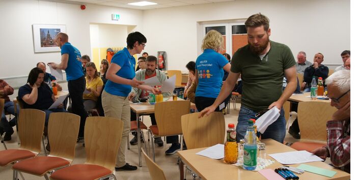 An mehreren Tischen sitzen in einem Raum viele Leute. Einige Personen gehen umher und Verteilen Erfassungsformulare, die von den Sitzenden ausgefüllt werden.