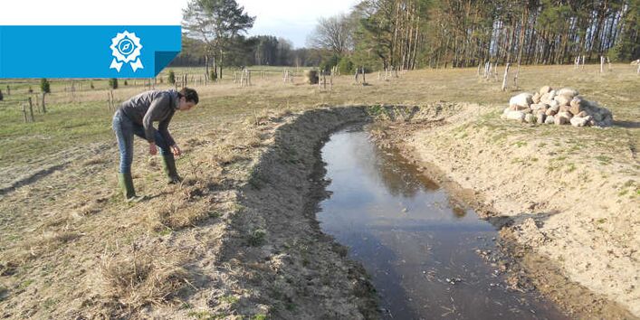 Eine Person beugt sich über einen Wassergraben, der durch ein Feld geht.