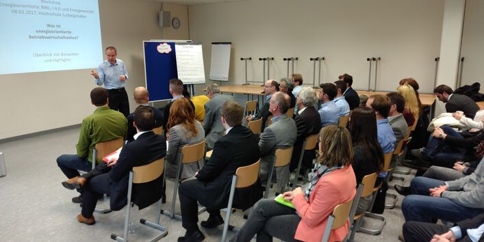 Votragender steht gestikulierend vor auf Stuhlreihen sitzendes Publikum. An seiner linken Seite befinden sich Flipcharts, Moderationswände und Tische. Im Hintergrund sieht man die Präsention auf der Wand.