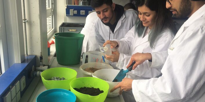 Mehrer Auszubildende hantieren mit verschiedenen Erden und Steinen in Schüssel. Eine Auszubildende gib Wasser mit einer Spritzflasche in eine der Schüsseln.