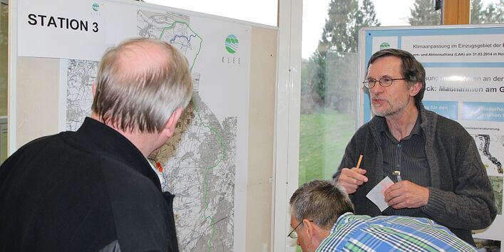 2 Männer stehen vor zwei Moderationswänden und sprechen miteinander. Auf den Moderationswänden sind Kartendarstellungen aufgepinnt. Ein Mann beugt sich näher herunter und betrachtet dabei genau die linke Karte. 