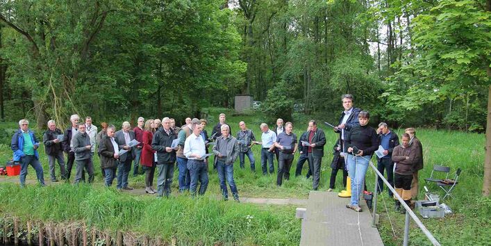 Personengruppe steht am Wasserlauf. Im Hintergrund stehen grün belaubte Bäumen. Eine Brücke führt über den Wasserlauf, auf der zwei Personen stehen.