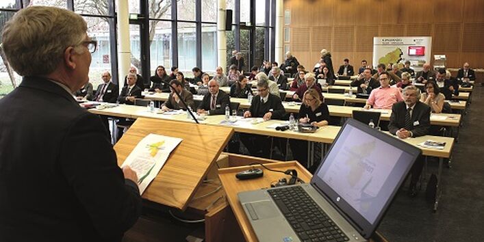 Vortragender steht am Pult bei geöffnetem Laptop vor den Teilnehmern, die in Reihen an Tischen sitzend zuhören.