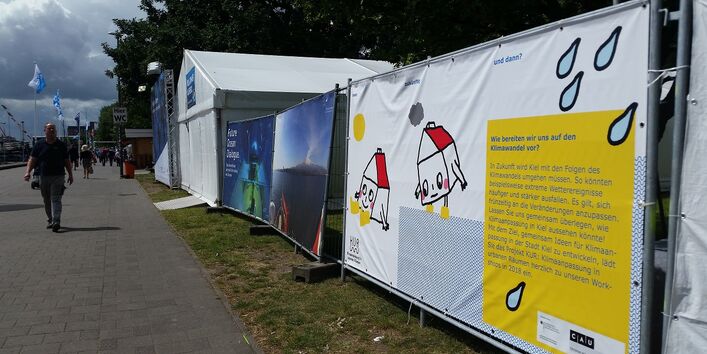 Das Projekt Banner sichtbar zur Straße hin auf der Kieler Woche 2017. Fußgänger laufen auf dem Gehweg. Auf dem Banner sind zwei Zeichnungen von Häusern mit Gesichtern zu sehen. Der Projekttext steht in einer gelben Textbox mit der Überschrift: Wie bereiten wir uns auf den Klimawandel vor.