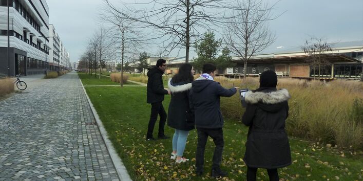 Auszubildende erkunden Maßnahmen zur Klimawandelanpassung vor Ort. Sie stehen auf dem Grünstreifen eines Gebäudeaußengeländes.