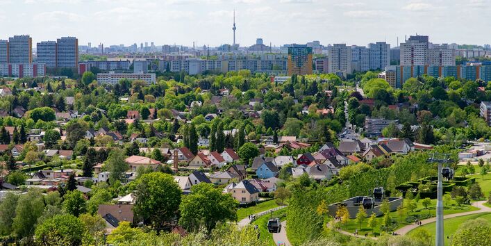 Vogelperspektive über einen dichten Berliner Park mit Seilbahn-Gondeln. Im Hintergrund die Skyline mit Fernsehturm und Wohngebäuden unter leicht bewölktem Himmel.