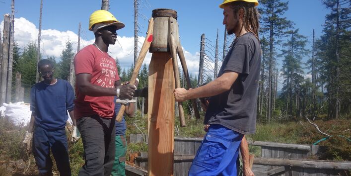 Zwei Männer stampfen einen Holzbalken in den Boden.