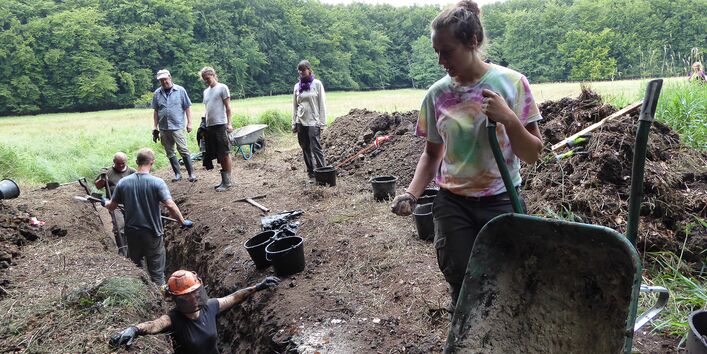 Mehrere Personen stehen auf dem umgegrabenen Boden einer Freifläche im Wald. Links im Bild ist ein Graben zu sehen, in dem mehrere Personen stehen und graben. Rechts im Vordergrund steht eine Person mit einer Schubkarre. Um den Graben herum stehen Eimer und eine weitere Schubkarre.