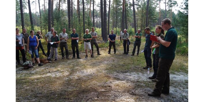 Eine Gruppen Menschen stehen im Halbkreis in einem Wald und verfolgen Erläuterungen einer Person. Viele halten Papier in den Händen.