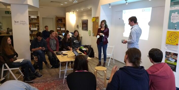 Vor einer Präsentation an der Wand stehen zwei Personen. Sie präsentieren mehreren anderen Personen, die im Halbkreis sitzen etwas.