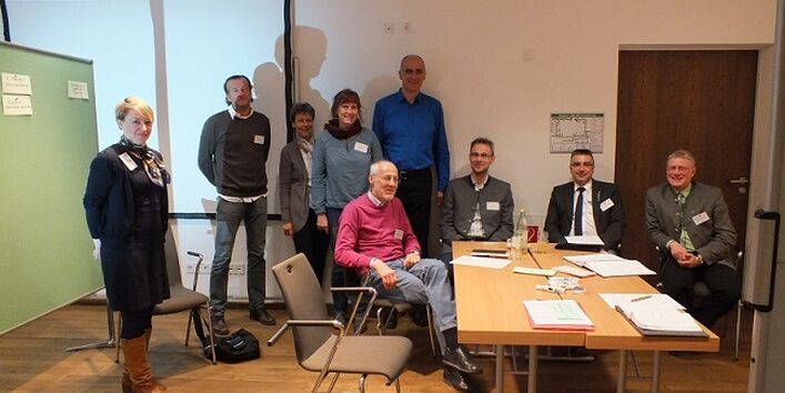 Workshopteilnehmerinnen und -teilnehmer sitzen am Arbeitsgruppentisch. Im Hintergrund stehen weitere Teilnehmerinnen und Teilnehmer vor Präsentationswänden.