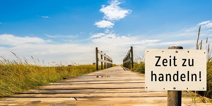 Auf einem Schild im Vordergrund steht Zeit zu handeln. Im Hintergrund führt ein Holzdünenweg optisch in den Himmel hinein.