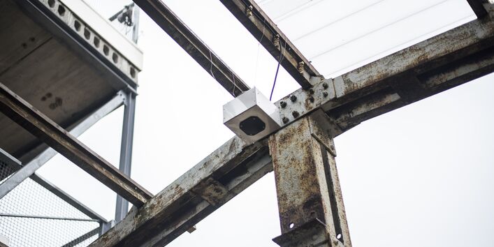 Dachkonstruktion aus Stahlträgern ohne Dachplatten; dazwischen hängt eine Metallbox, die Regenwasserskulptur.