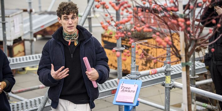 Rechts ein Zierapfelbaum mit vielen Früchten, in der Mitte ein Schild "Tree Aware", links ein Mensch im Gespräch.