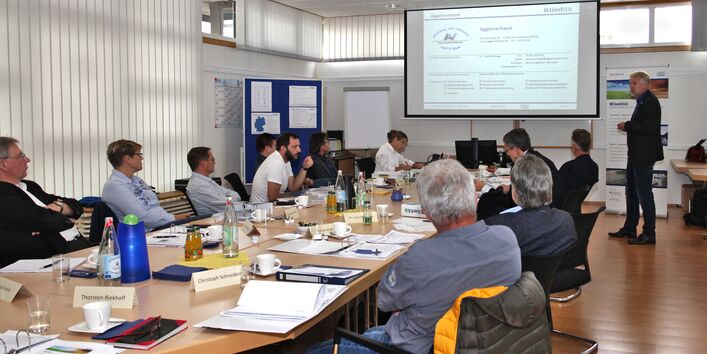 In einem großen Raum sitzen mehrere Personen an einem langen Konferenztisch. Am Kopfende des Tisches wird auf eine Leinwand eine Präsentation projiziert, daneben steht ein Mann und hält einen Vortrag.