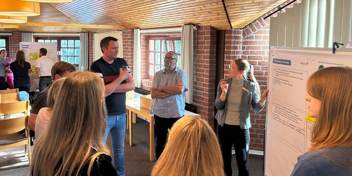 Personen stehen im Raum verteilt vor Flipcharts mit Texten und Grafiken. Der Raum hat Holzdecken und besteht aus Klinkerbauweise