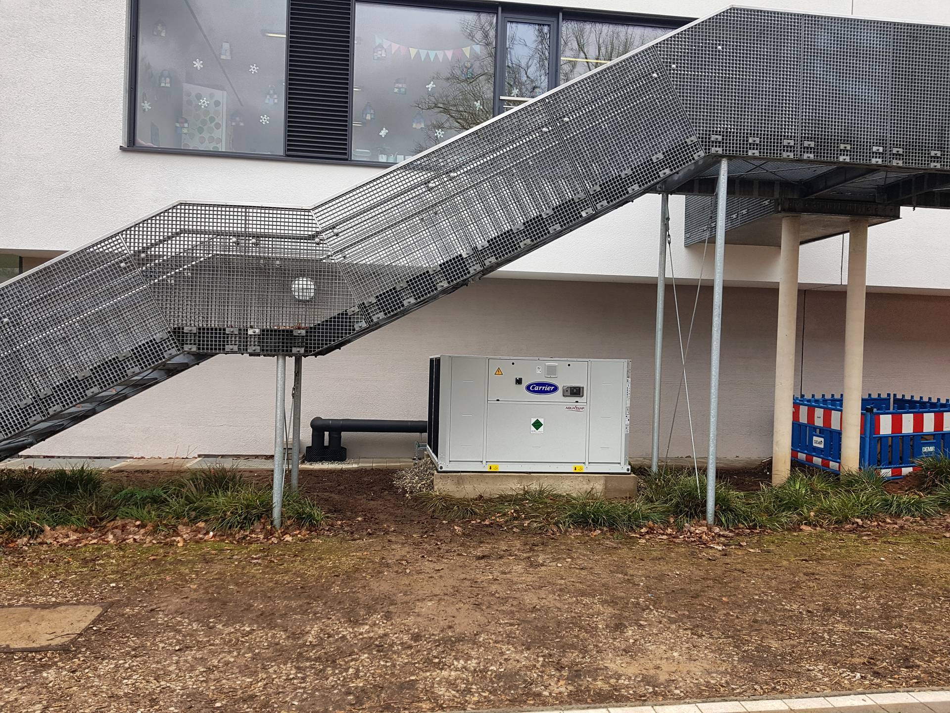 Eine große Wärmepumpe ist an der Fassade eines Schulgebäudes unter einer Treppe platziert.