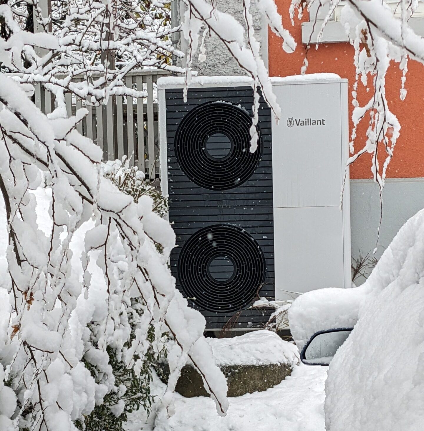 Außengerät einer Wärmepumpe vor einem Gebäude. Im Gerät zwei Lüfter, auf dem Gerät und in der Umgebung liegt 10 cm Schnee.
