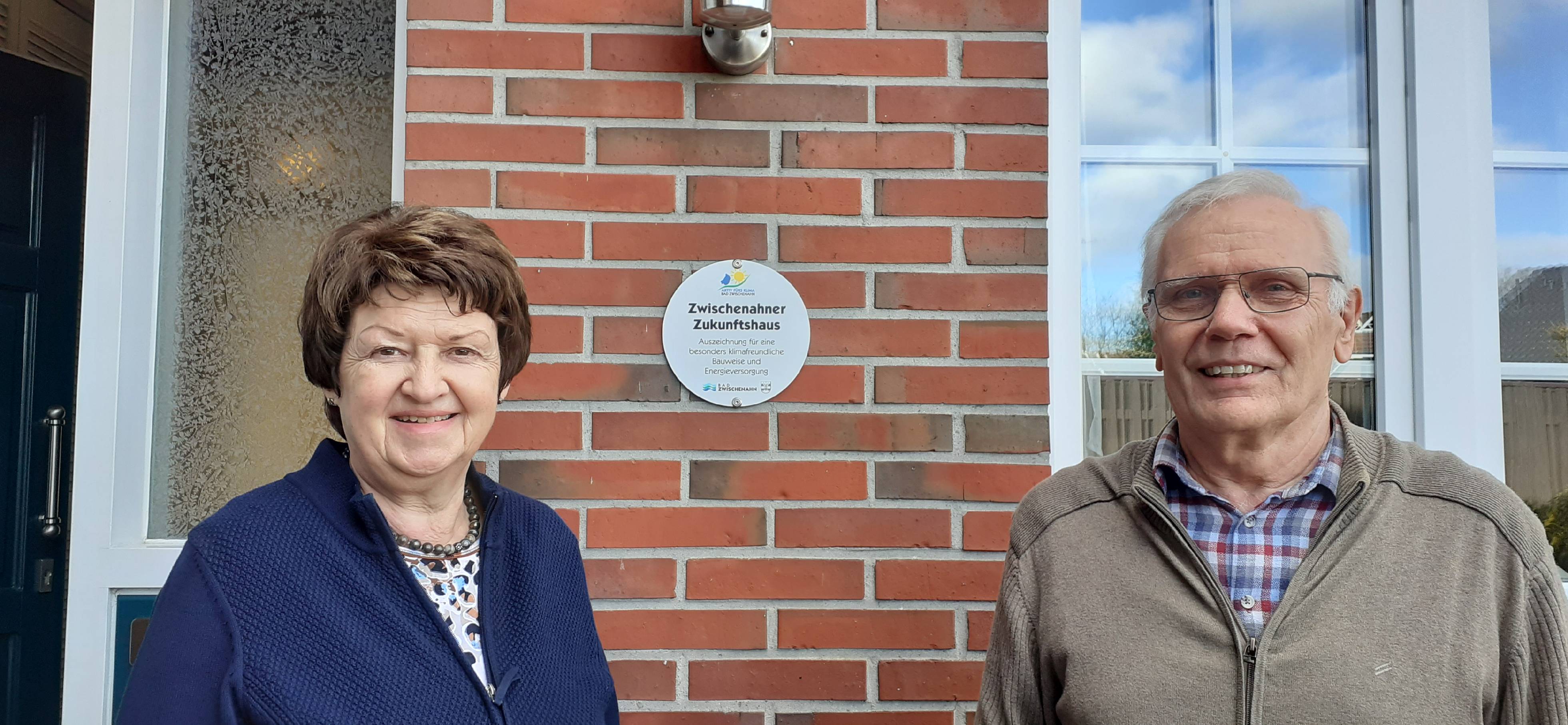 Die zwei Hausbewohner vor ihrem Haus mit einer Plakette für die Auszeichnung als Zukunftshaus an der Wand.